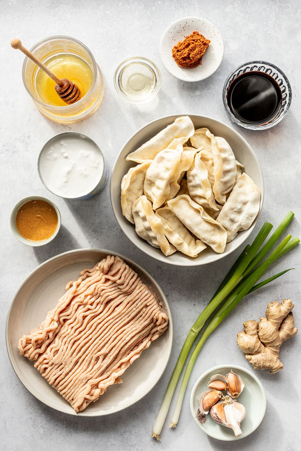 Assorted ingredients on a countertop including raw dumplings, ground meat, green onions, ginger, garlic, soy sauce, honey, miso paste, and a small bowl of white liquid.