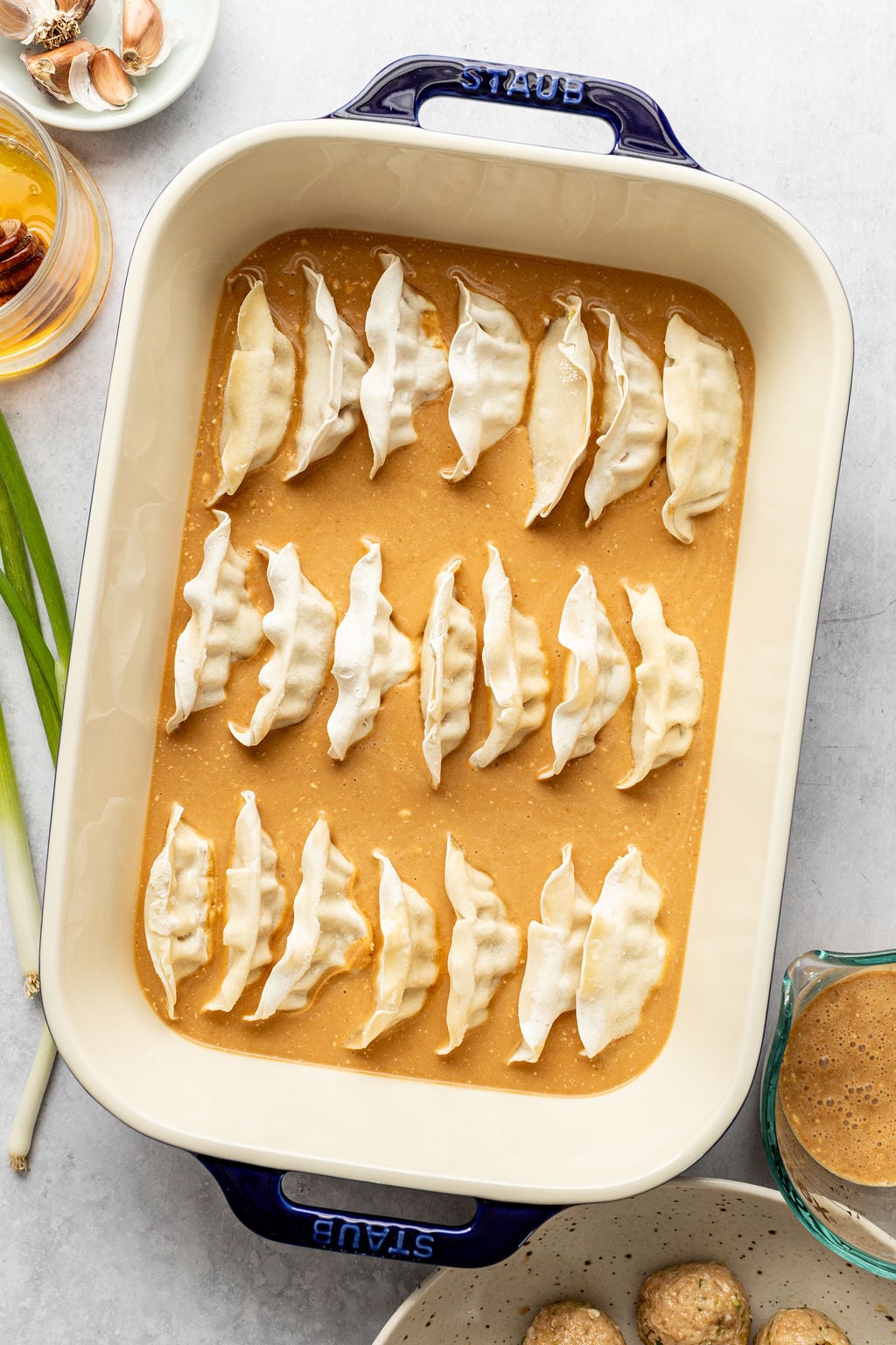 Uncooked dumplings arranged in rows in a creamy orange sauce inside a rectangular baking dish, surrounded by ingredients on a countertop.