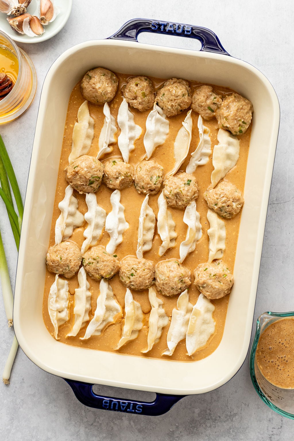A baking dish filled with raw meatballs and uncooked dumplings arranged in a light brown sauce, ready to be cooked.