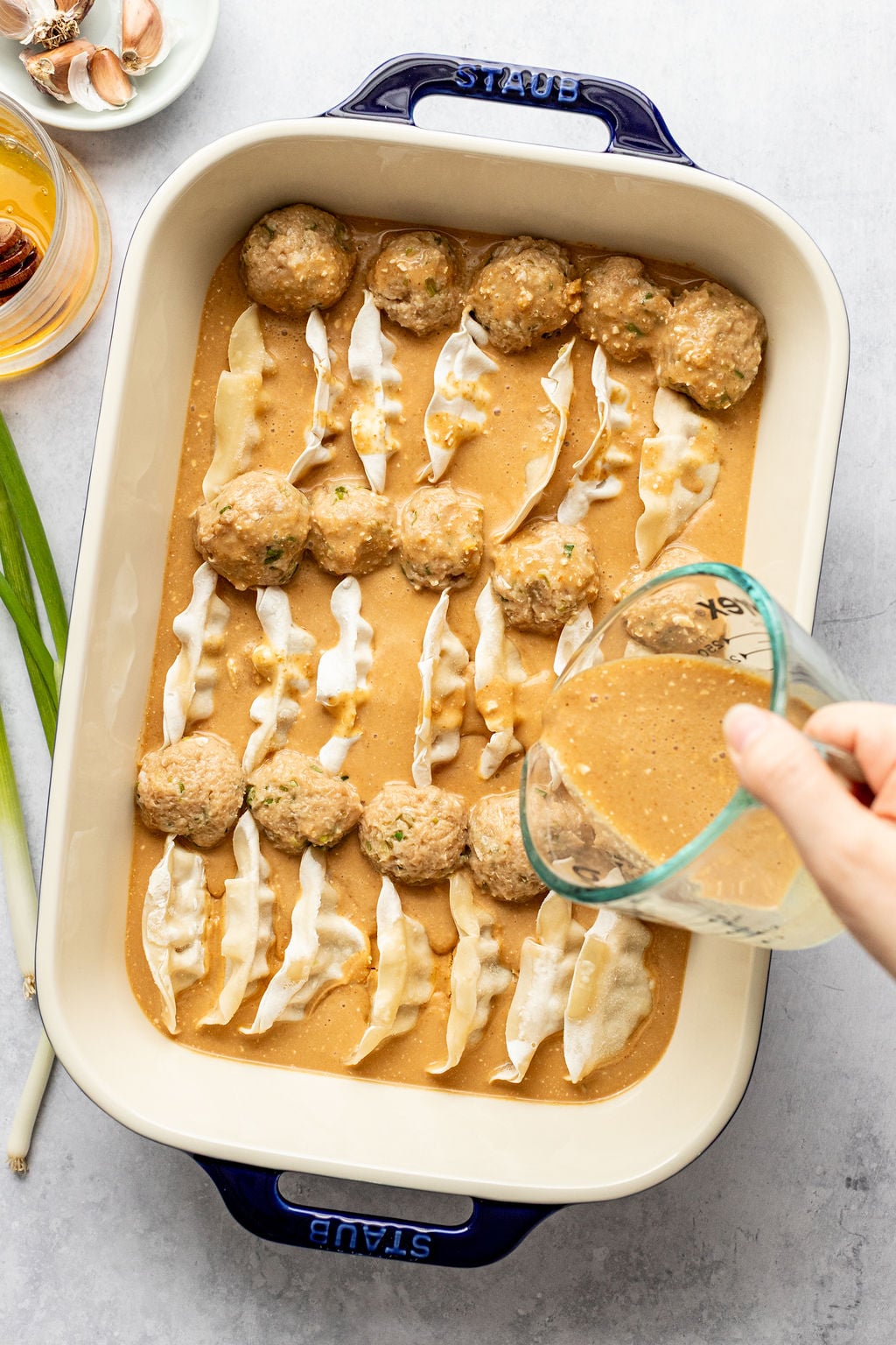 A hand pours sauce over rows of dumplings and meatballs in a rectangular baking dish on a light surface, with ingredients visible nearby.