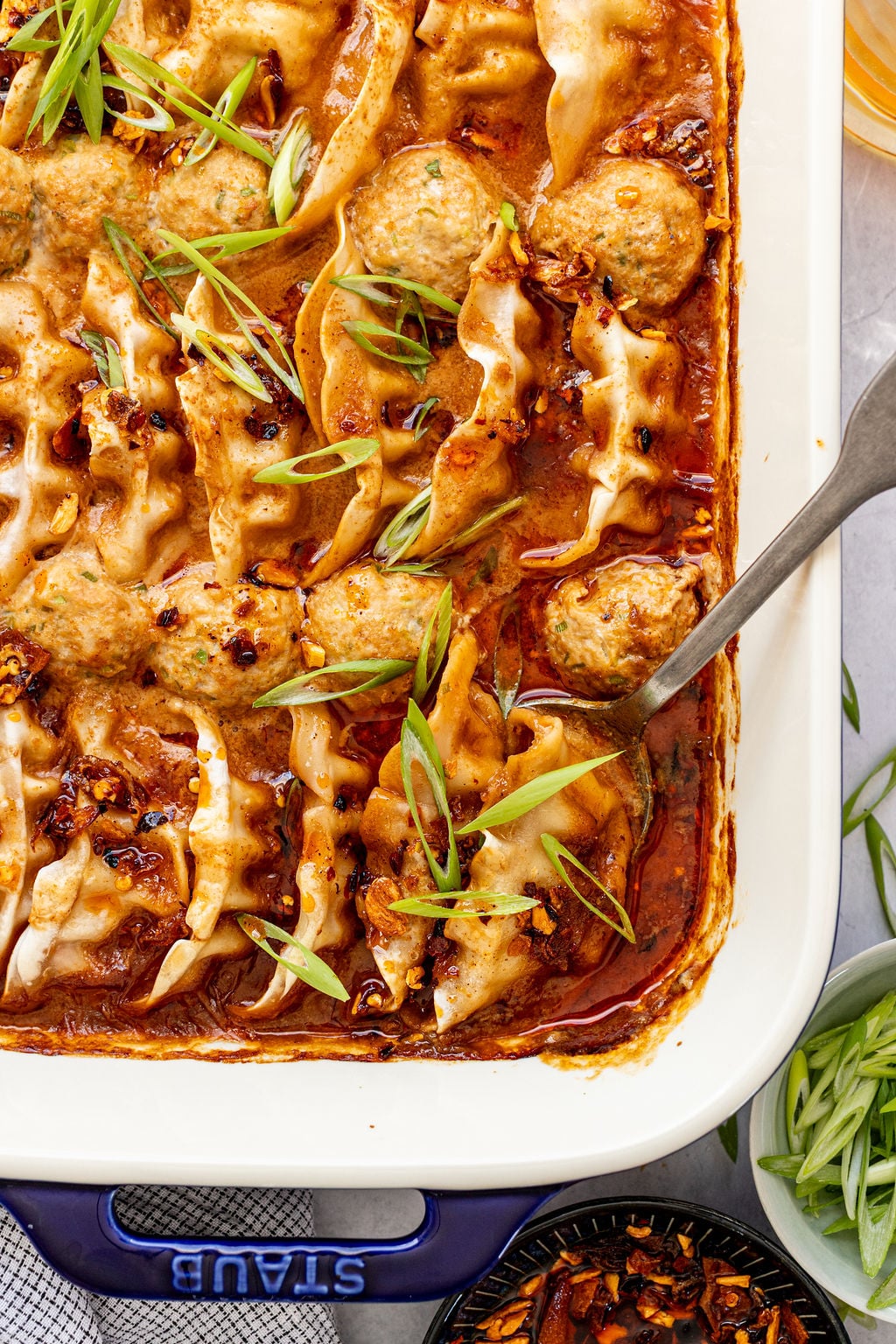 Baked dumplings in a red sauce topped with sliced green onions, served in a rectangular Staub dish with a metal serving spoon.