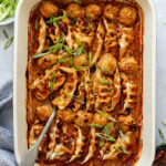 A baking dish filled with dumplings and meatballs in a red sauce, garnished with sliced green onions, with a serving spoon on the side.