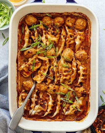 A baking dish filled with dumplings and meatballs in a red sauce, garnished with sliced green onions, with a serving spoon on the side.