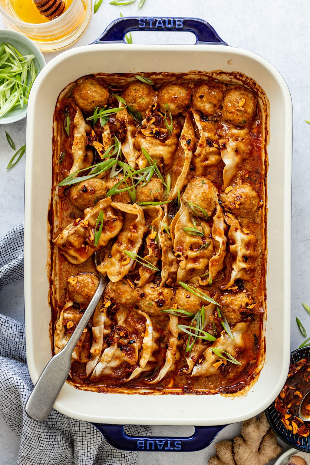 A baking dish filled with dumplings and meatballs in a red sauce, garnished with sliced green onions, with a serving spoon on the side.