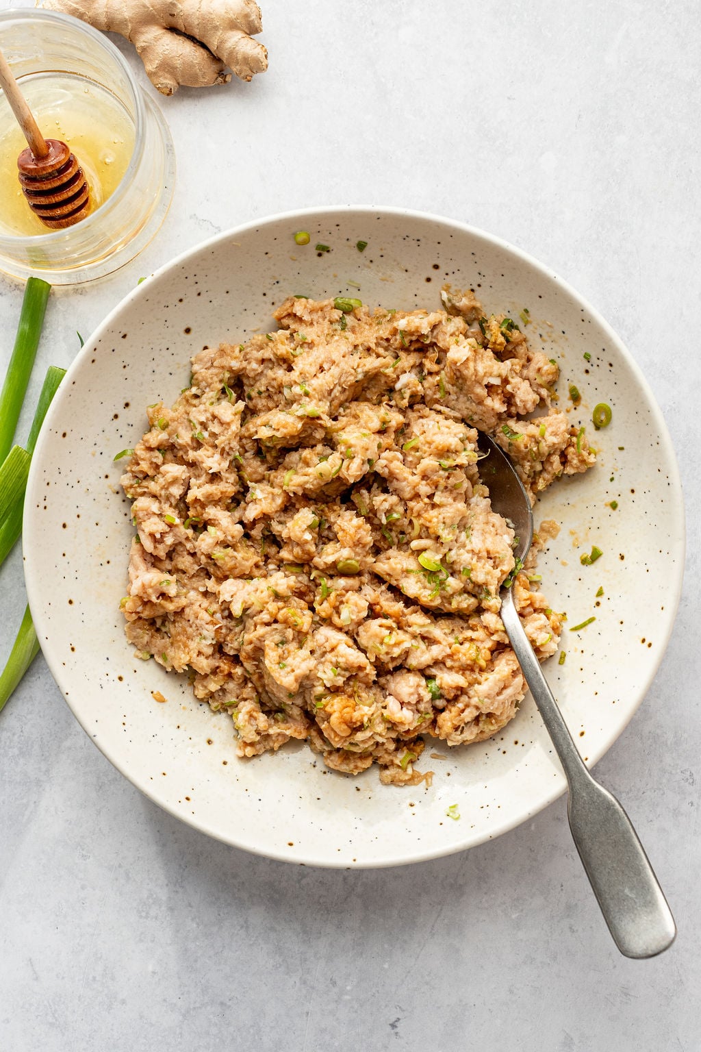 A bowl of seasoned minced meat mixture with a spoon, next to a jar of honey, ginger root, and a green onion on a light surface.