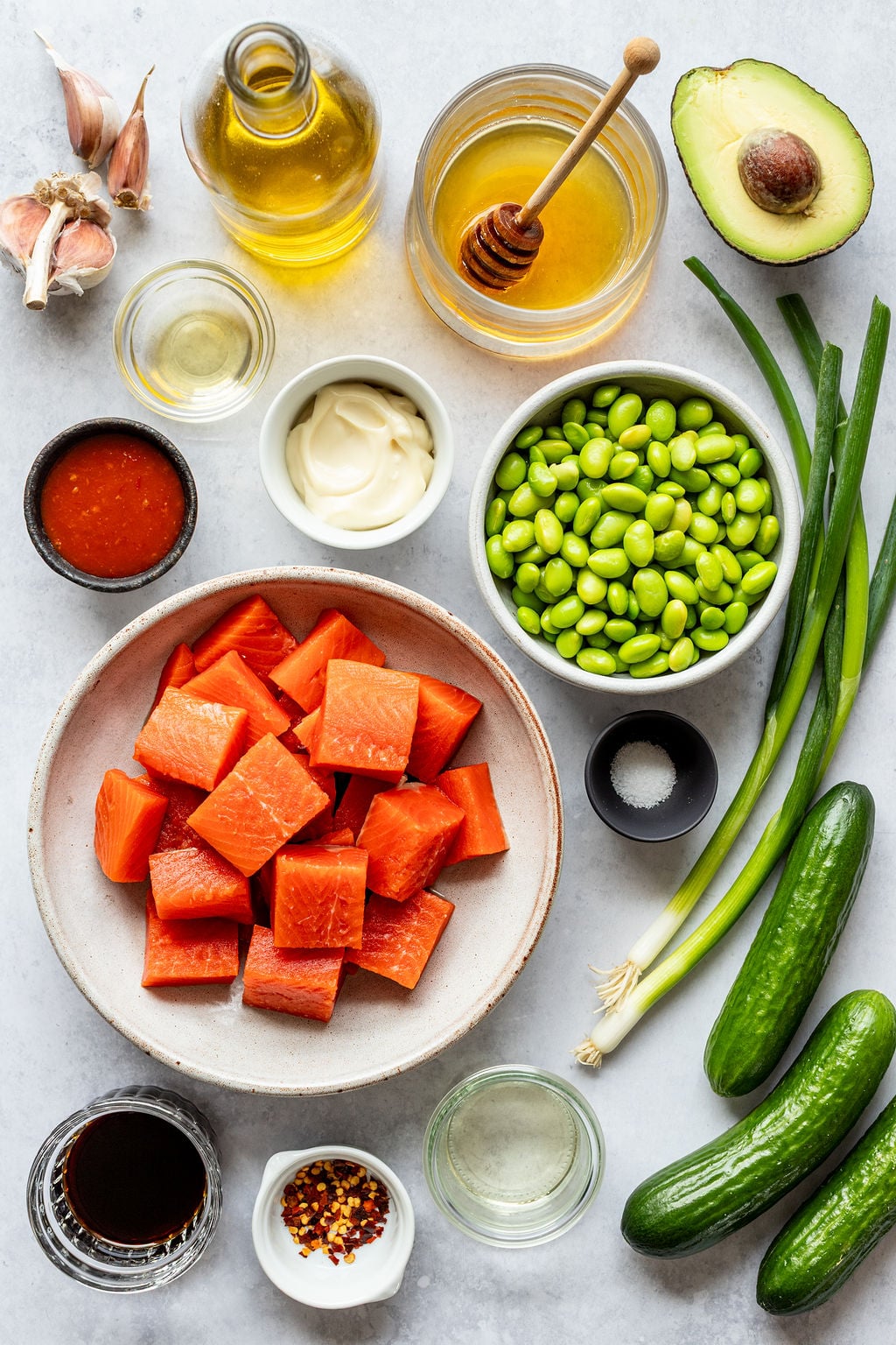 Various ingredients for a salmon bowl arranged on a surface, including salmon cubes, edamame, cucumber, avocado, sauces, seasonings, olive oil, honey, garlic, and green onions.