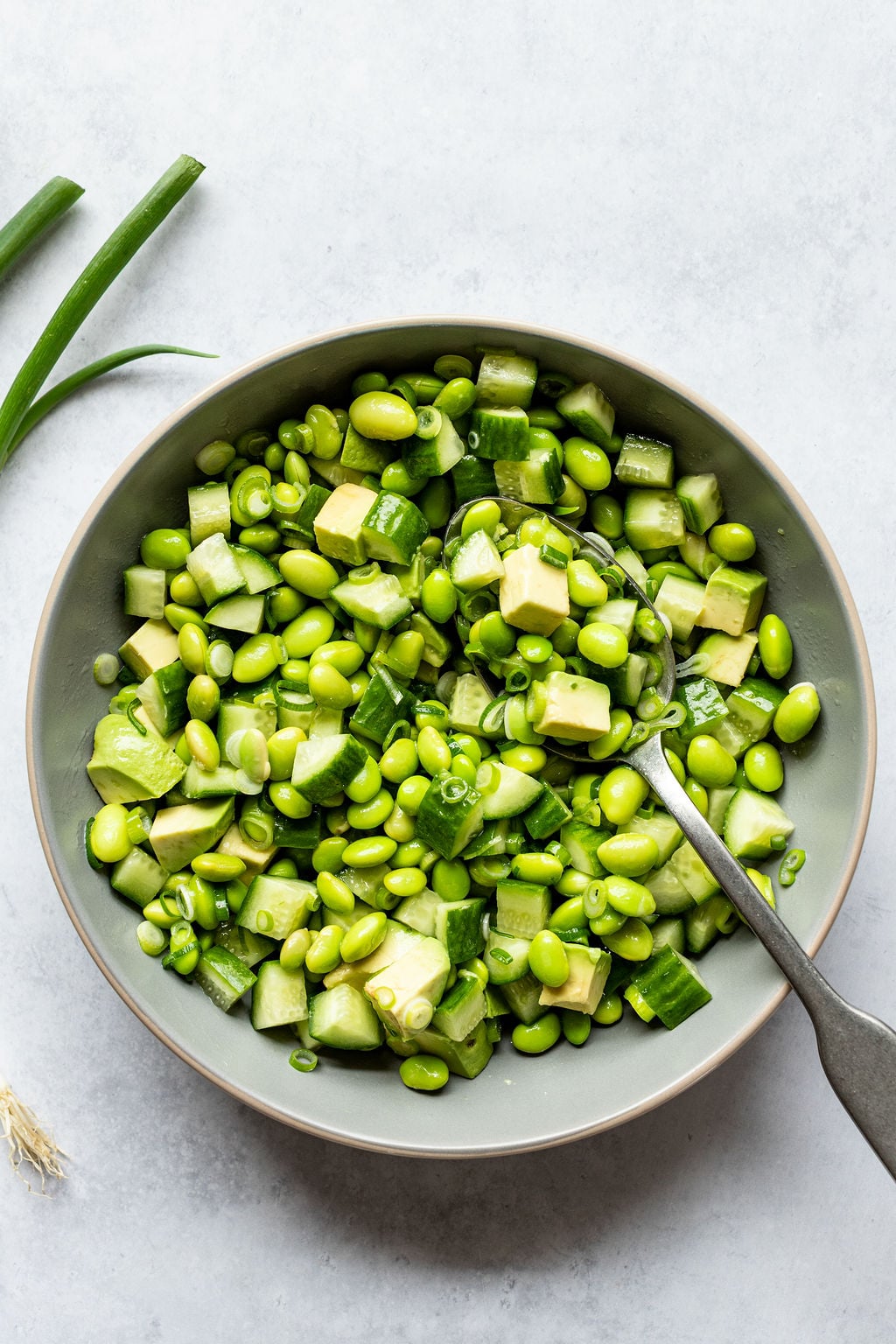 A bowl of salad containing chopped cucumber, avocado, edamame, and green onions, with a spoon resting in the bowl on a light background.