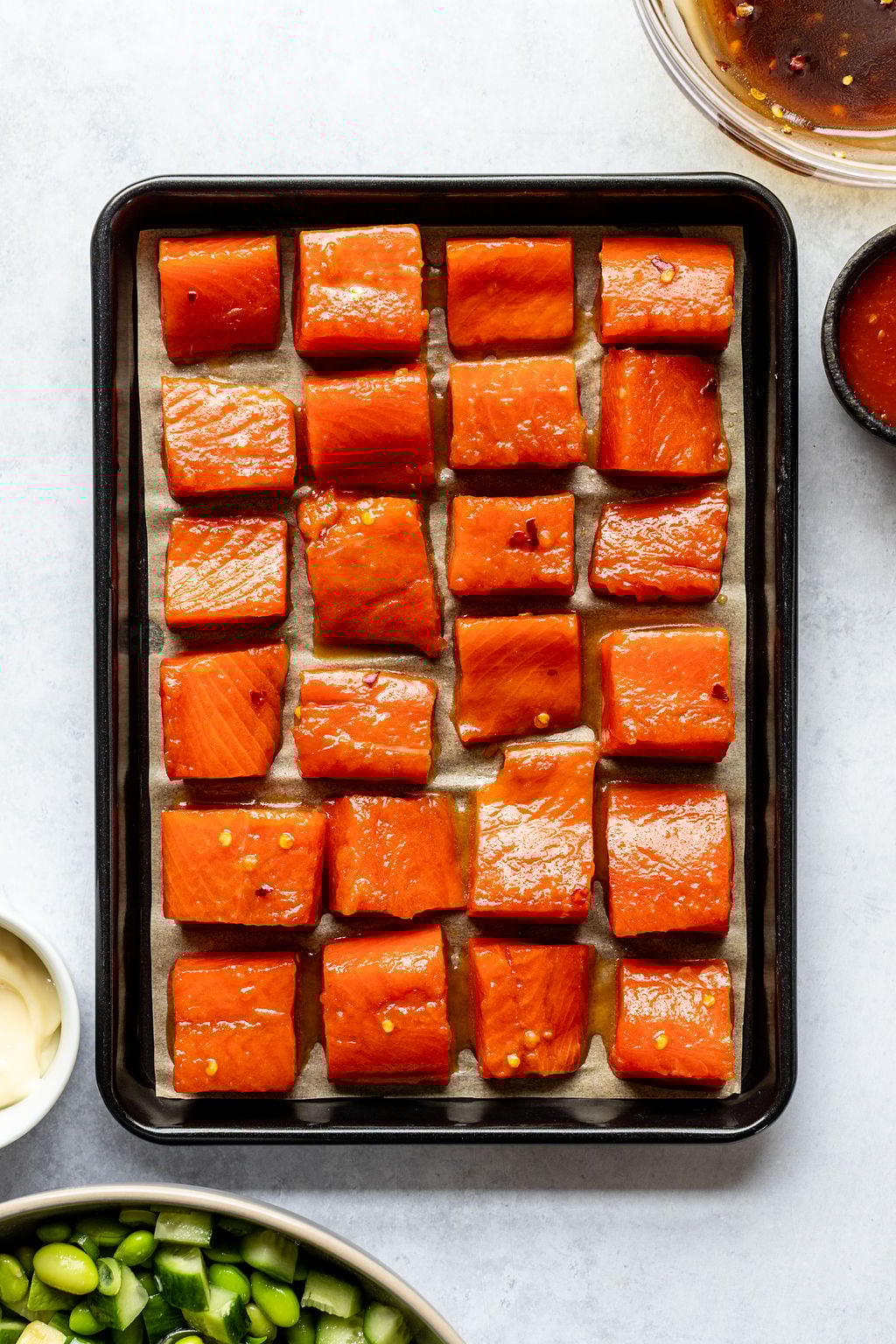 A baking tray lined with parchment paper holds evenly spaced cubes of salmon coated in a red marinade, surrounded by bowls of sauce and chopped vegetables.