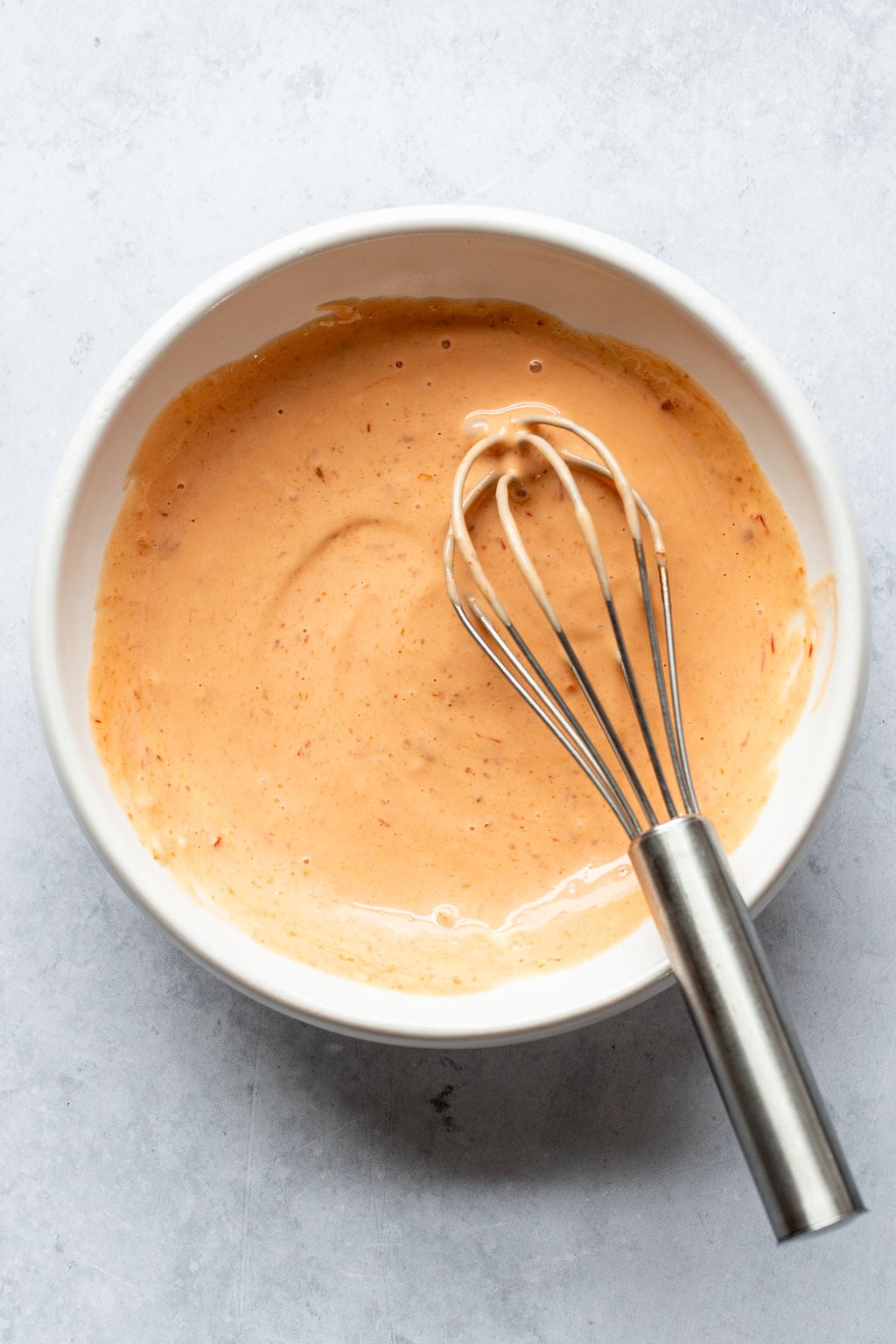 A metal whisk rests in a white bowl filled with a creamy orange sauce, placed on a light gray surface.