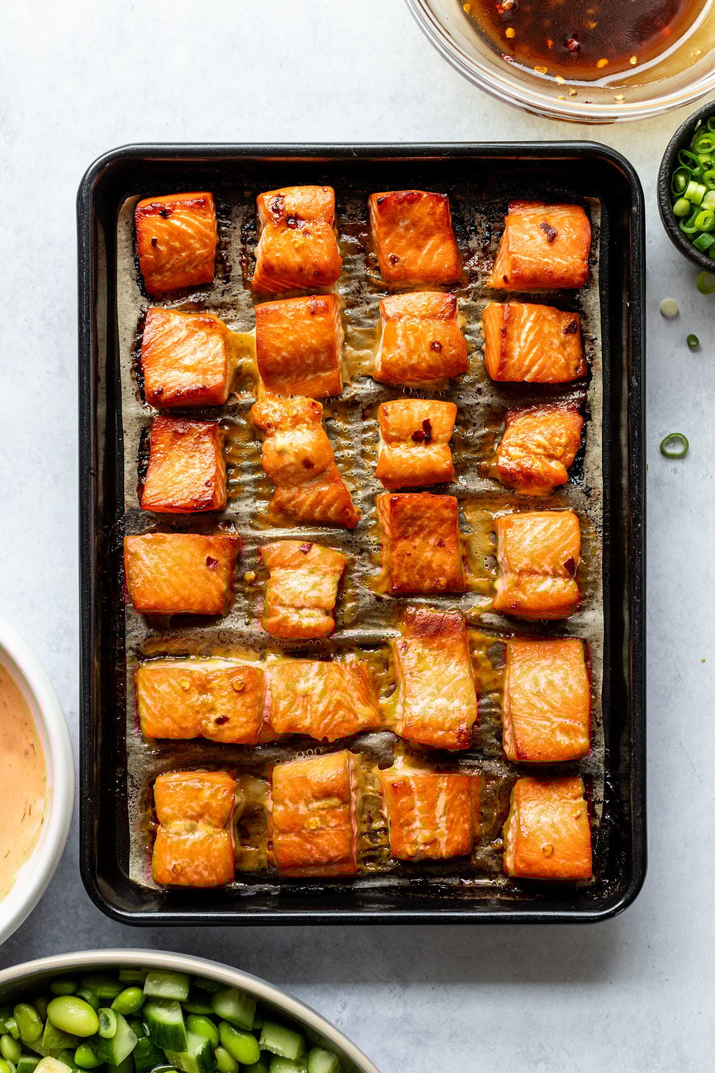 A baking tray with evenly spaced cooked salmon cubes. Surrounding the tray are bowls of sauce and chopped green onions.