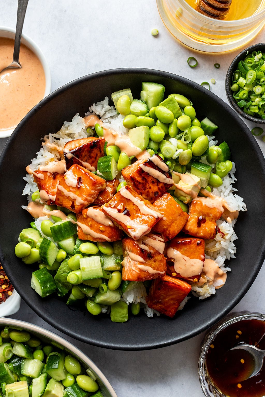 A bowl of rice topped with glazed salmon, diced cucumber, edamame, green onions, and drizzled with creamy sauce; surrounded by small bowls of sauce and ingredients.
