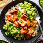 A black bowl containing white rice, glazed salmon pieces, edamame, sliced cucumber, and chopped green onions, with a fork on the side.
