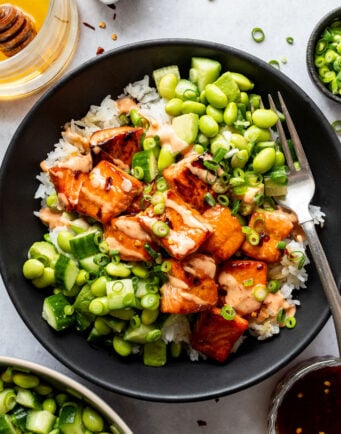 A black bowl containing white rice, glazed salmon pieces, edamame, sliced cucumber, and chopped green onions, with a fork on the side.