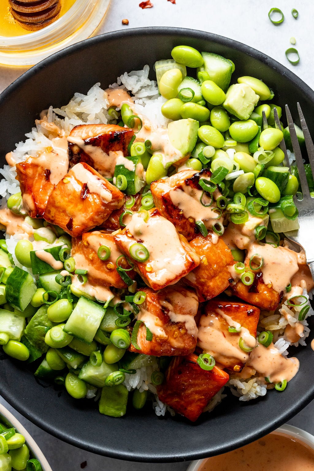 A bowl of white rice topped with glazed salmon cubes, sliced cucumbers, edamame, chopped green onions, and a creamy sauce.