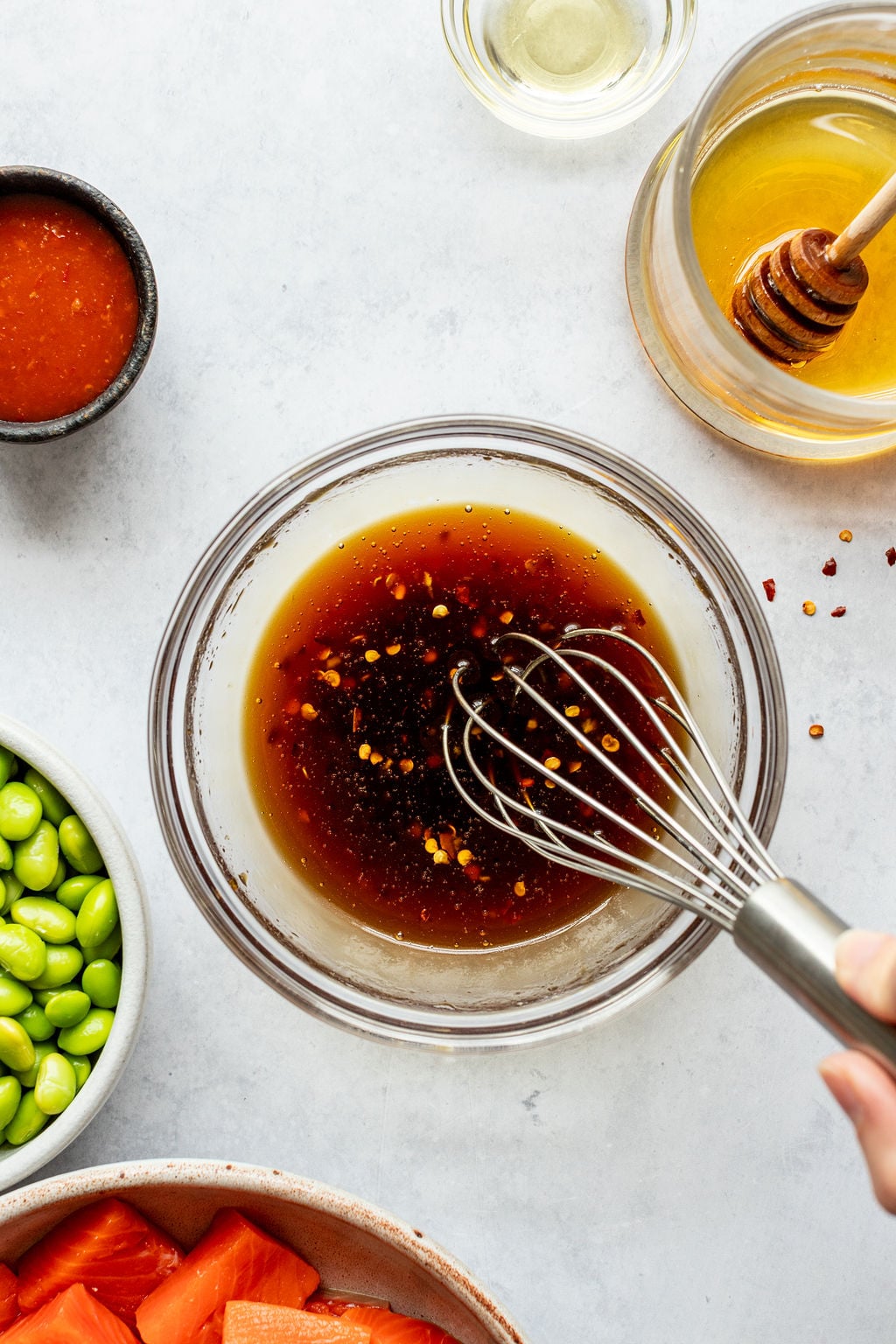 A hand whisking a brown sauce with chili flakes in a glass bowl, surrounded by small bowls of honey, oil, hot sauce, edamame, and sliced salmon cubes.