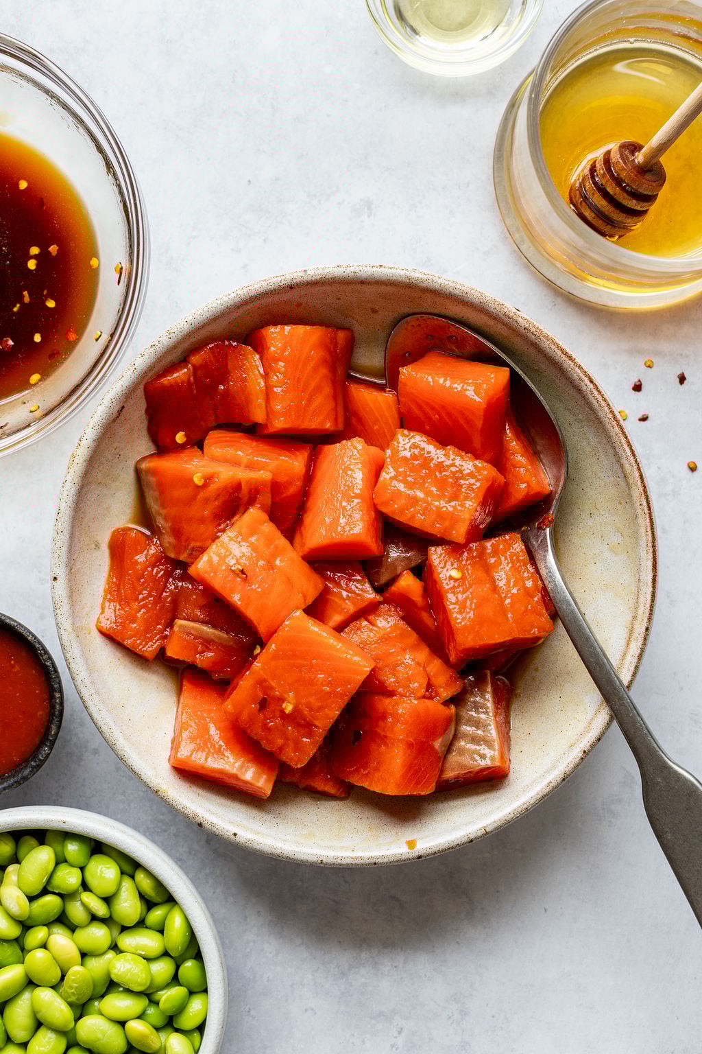 A bowl of cubed raw salmon with a spoon, surrounded by small bowls of edamame, sauces, and a jar of honey with a dipper.