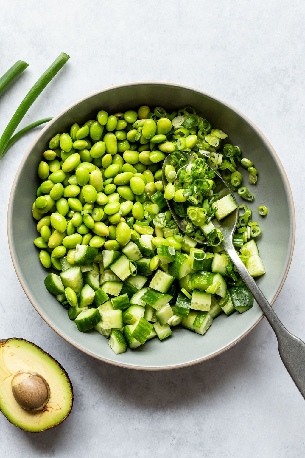 A bowl containing chopped cucumber, sliced green onions, and shelled edamame, with a spoon inside. An avocado half and green onions are beside the bowl on a white surface.