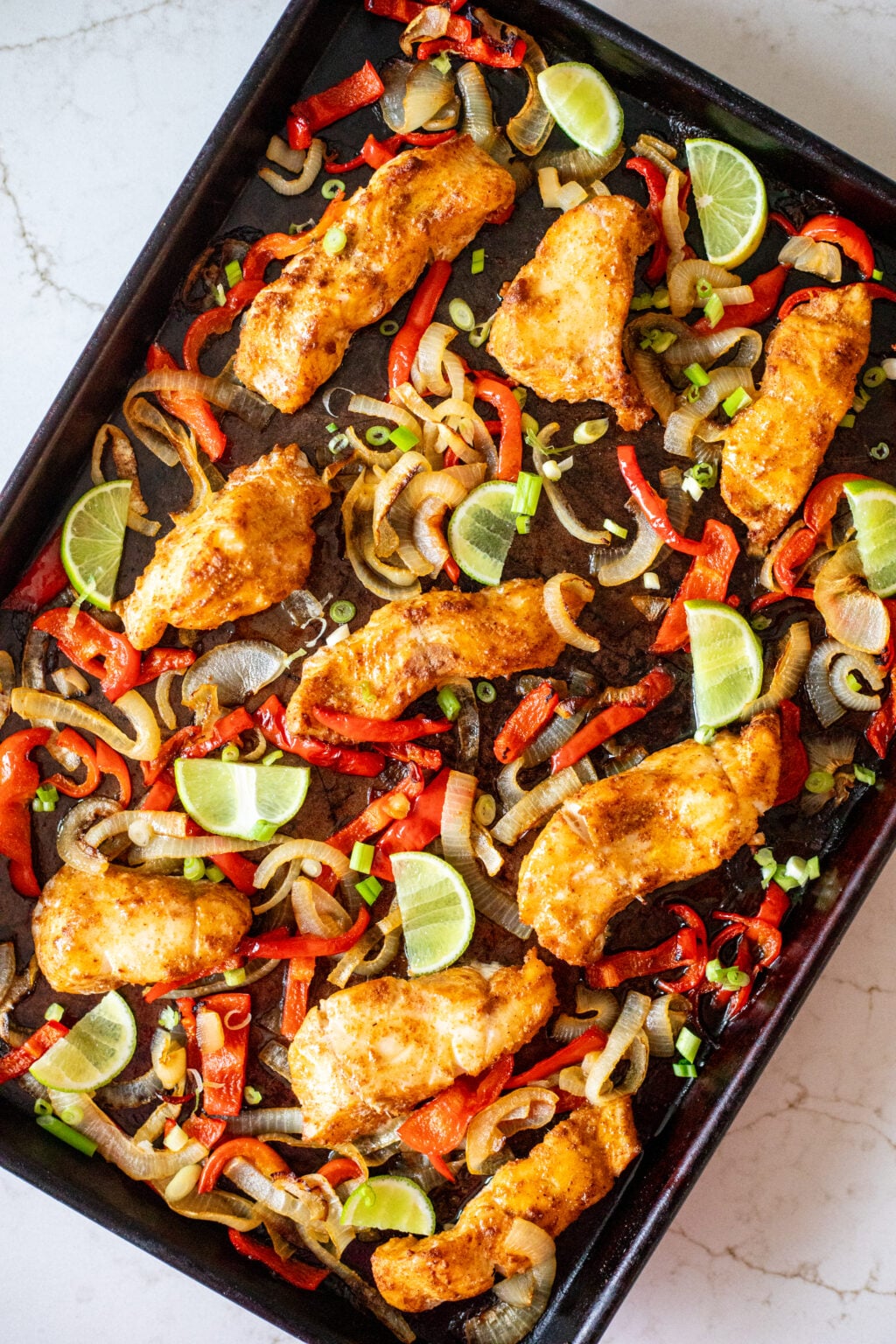 Roasted spiced white fish with peppers, onions, and lime slices on a black sheet pan.