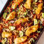 Roasted spiced white fish with peppers, onions, and lime slices on a black sheet pan.