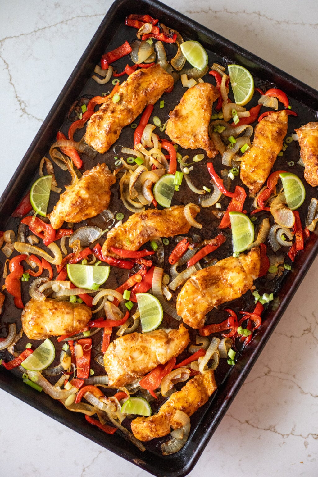 Roasted spiced white fish with peppers, onions, and lime slices on a black sheet pan.