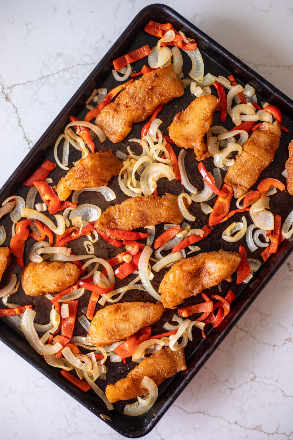 Spiced fish, onions, and peppers on a black sheet pan.