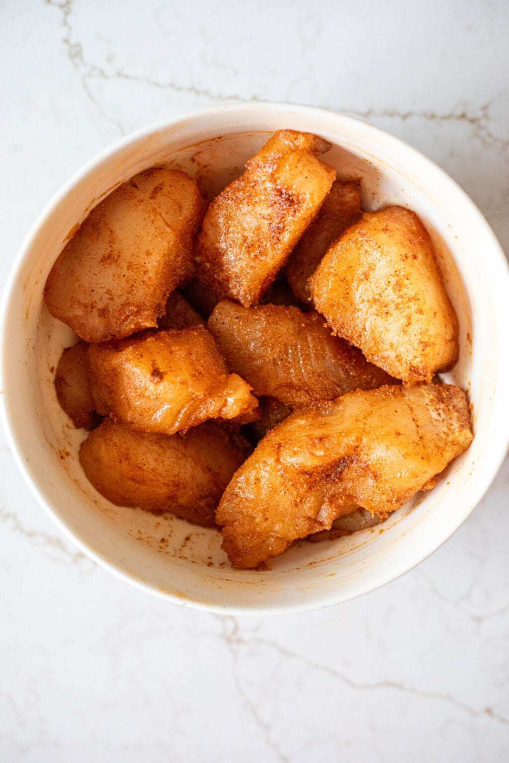 Bowl containing raw fish pieces coated with a reddish seasoning mixture, placed on a white marble surface.