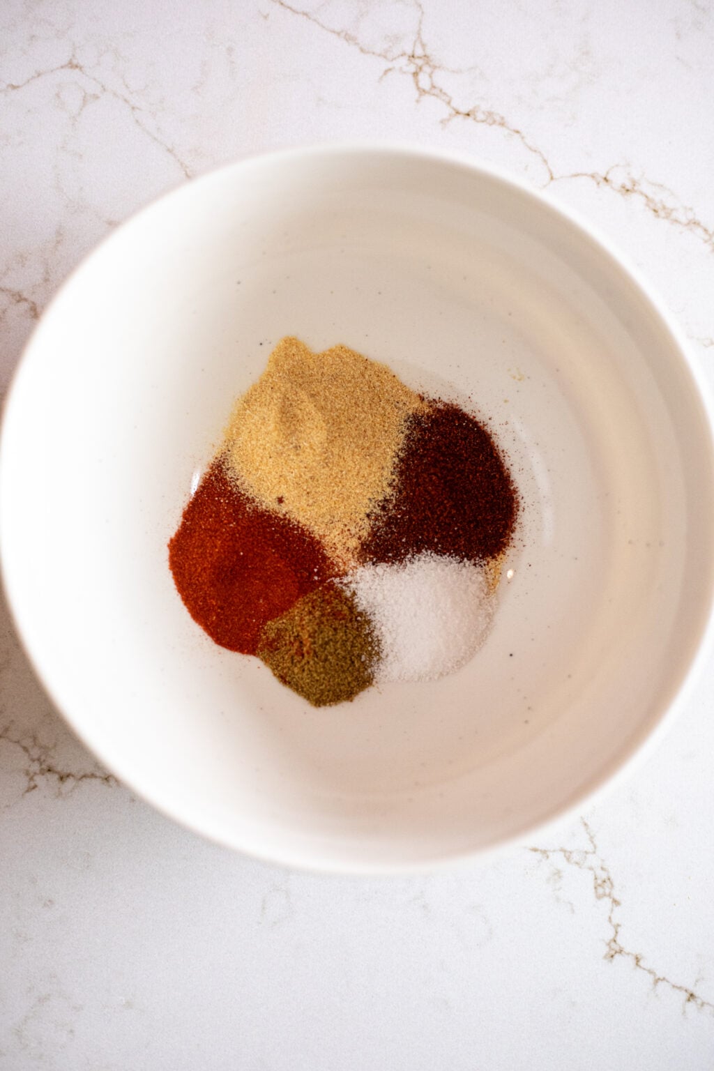 A white bowl with small piles of various ground spices and salt on a white marble surface.