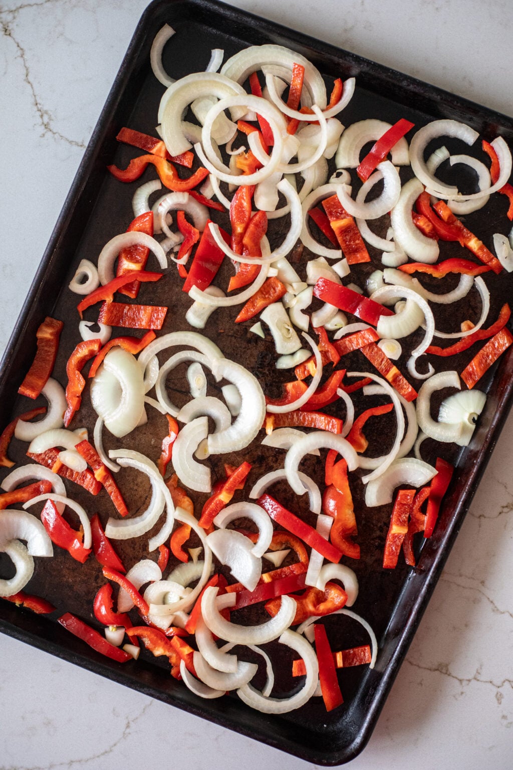 Sliced onions and red bell peppers spread on a dark baking sheet, ready for roasting on a light marbled countertop.
