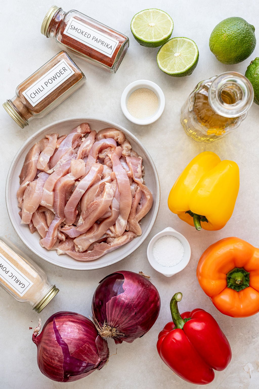Raw chicken strips on a plate surrounded by red, yellow, and orange bell peppers, red onions, lime halves, spices in jars, a jar of oil, and small bowls of seasonings.