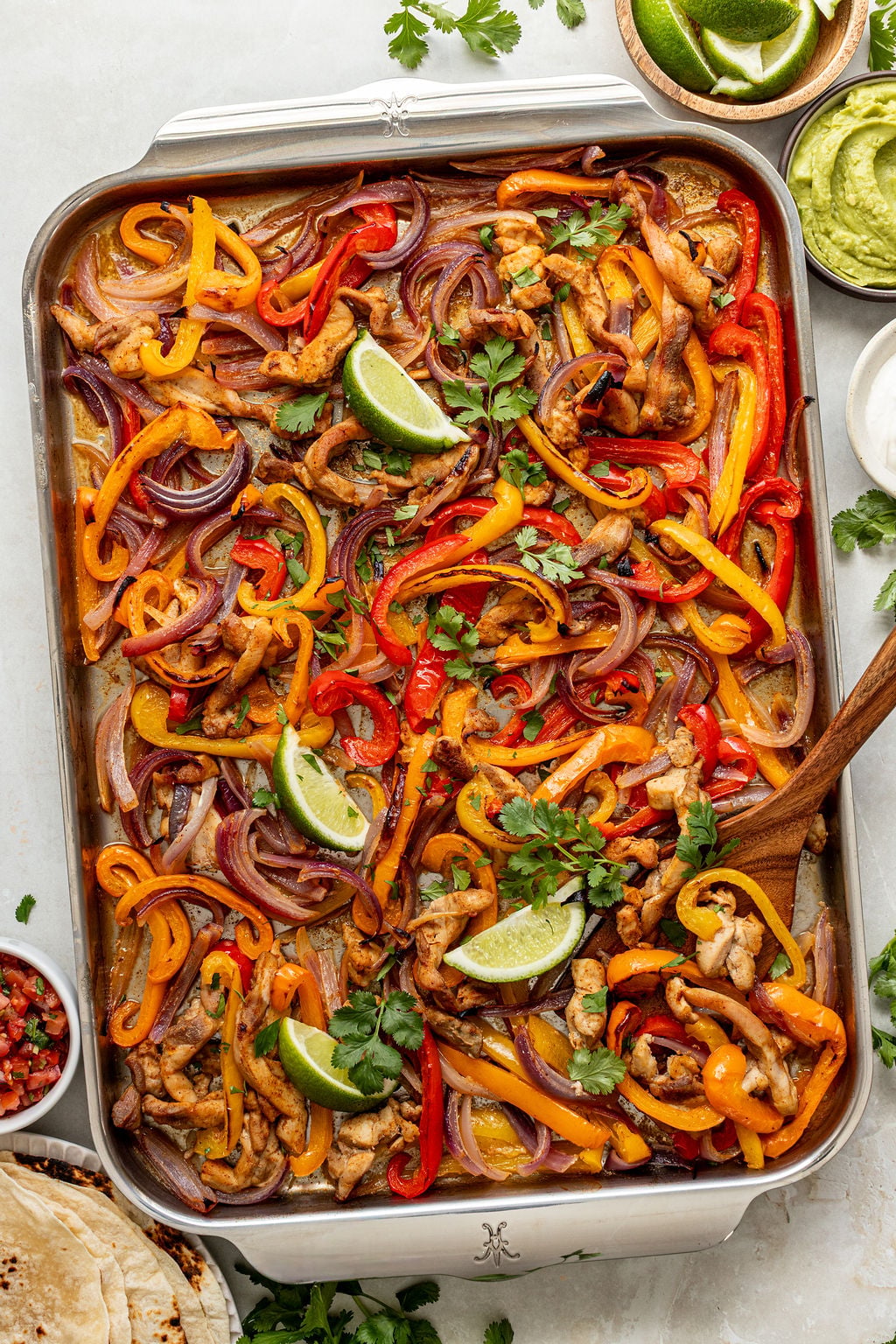 Sheet pan chicken fajitas with sliced bell peppers and onions, garnished with lime wedges and cilantro, served with sides of guacamole, salsa, tortillas, and lime wedges.