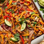 Sheet pan chicken fajitas with sliced bell peppers, red onions, and lime wedges, garnished with cilantro; served with guacamole, pico de gallo, and tortillas on the side.