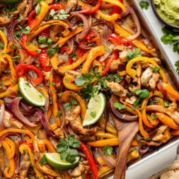 Sheet pan chicken fajitas with sliced bell peppers, red onions, and lime wedges, garnished with cilantro; served with guacamole, pico de gallo, and tortillas on the side.