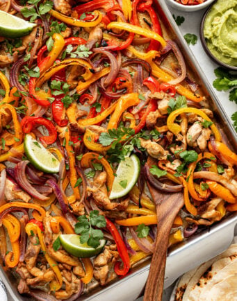 Sheet pan chicken fajitas with sliced bell peppers, red onions, and lime wedges, garnished with cilantro; served with guacamole, pico de gallo, and tortillas on the side.