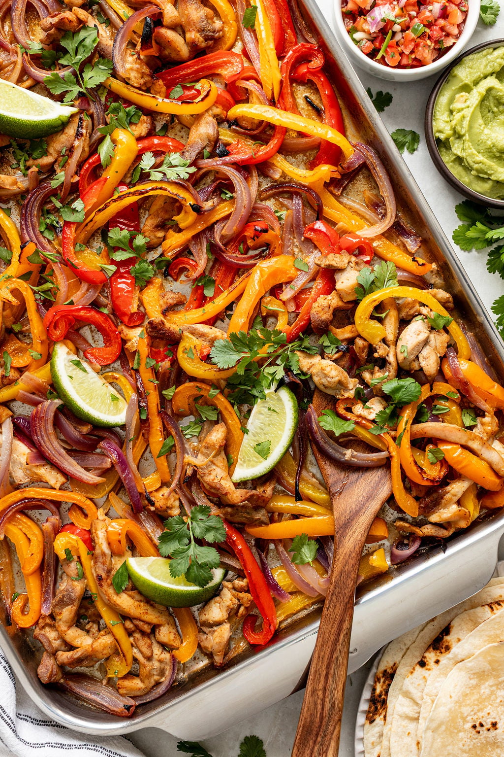 Sheet pan chicken fajitas with sliced bell peppers, red onions, and lime wedges, garnished with cilantro; served with guacamole, pico de gallo, and tortillas on the side.