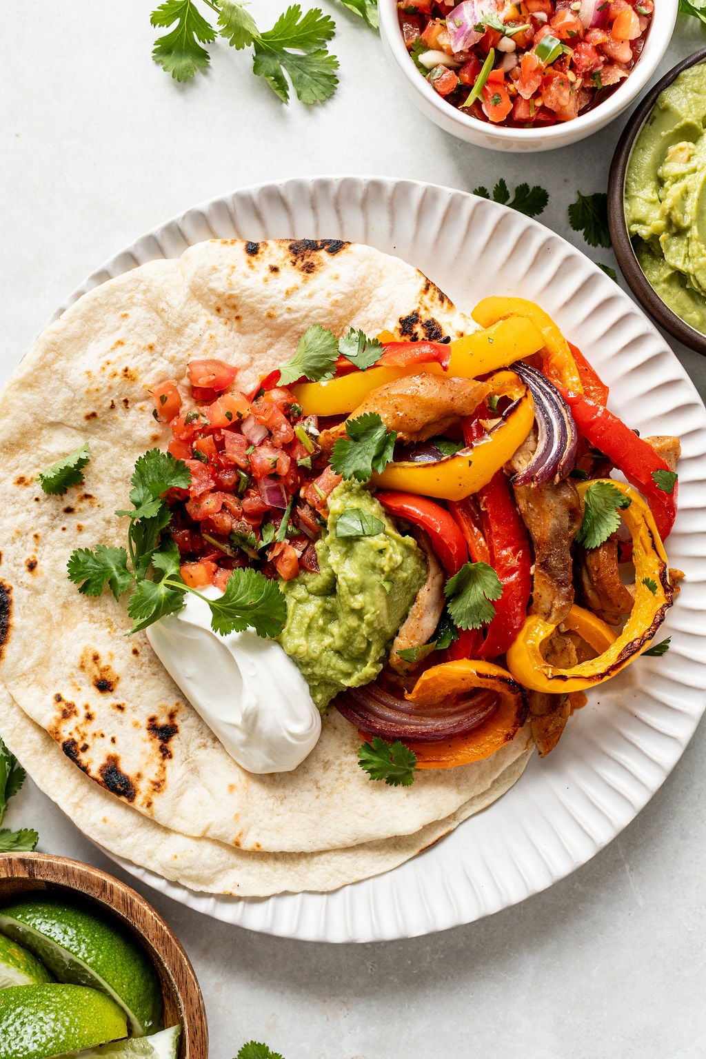 A plate with a tortilla topped with grilled peppers, onions, chicken, pico de gallo, guacamole, sour cream, and cilantro.