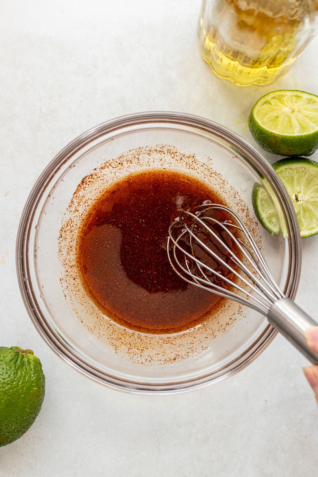 A hand whisking a reddish-brown liquid in a glass bowl, with halved limes, a whole avocado, and a glass of light-colored liquid nearby on a white surface.
