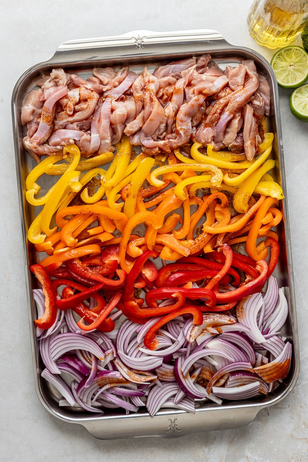 Sheet pan with rows of raw sliced chicken, yellow, orange, and red bell peppers, and red onions, arranged in sections and seasoned.