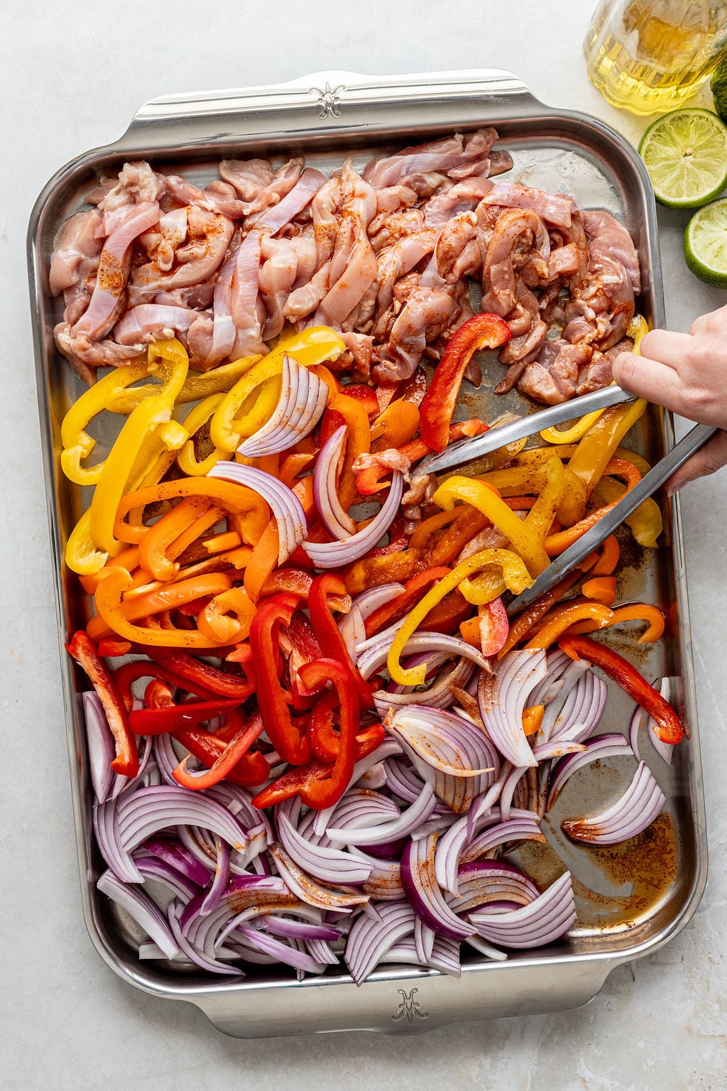 A tray with sliced raw chicken, red onions, and red, yellow, and orange bell peppers; a hand uses tongs to mix the vegetables.