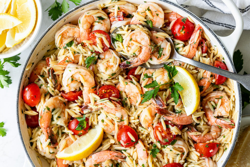 A skillet filled with shrimp, orzo pasta, cherry tomatoes, fresh parsley, and lemon wedges, with a serving spoon and lemon slices on the side.