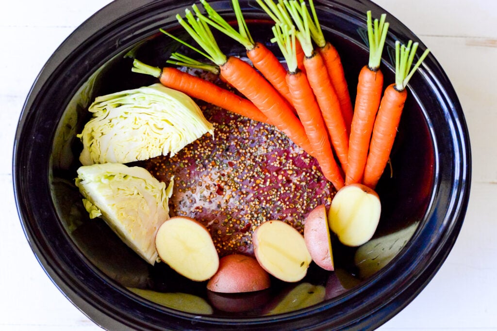Crockpot filled with uncooked corned beef, halved red potatoes, sliced cabbage, and whole carrots with tops, sprinkled with spices.