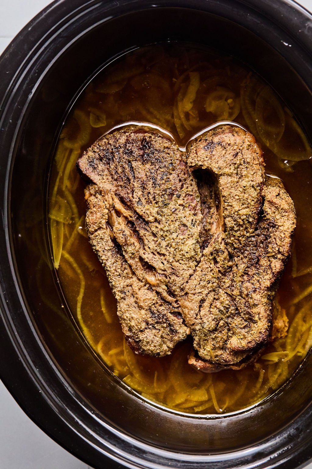 Two seasoned beef chuck roasts that are cooking in broth inside a slow cooker, with sliced onions visible in the liquid.