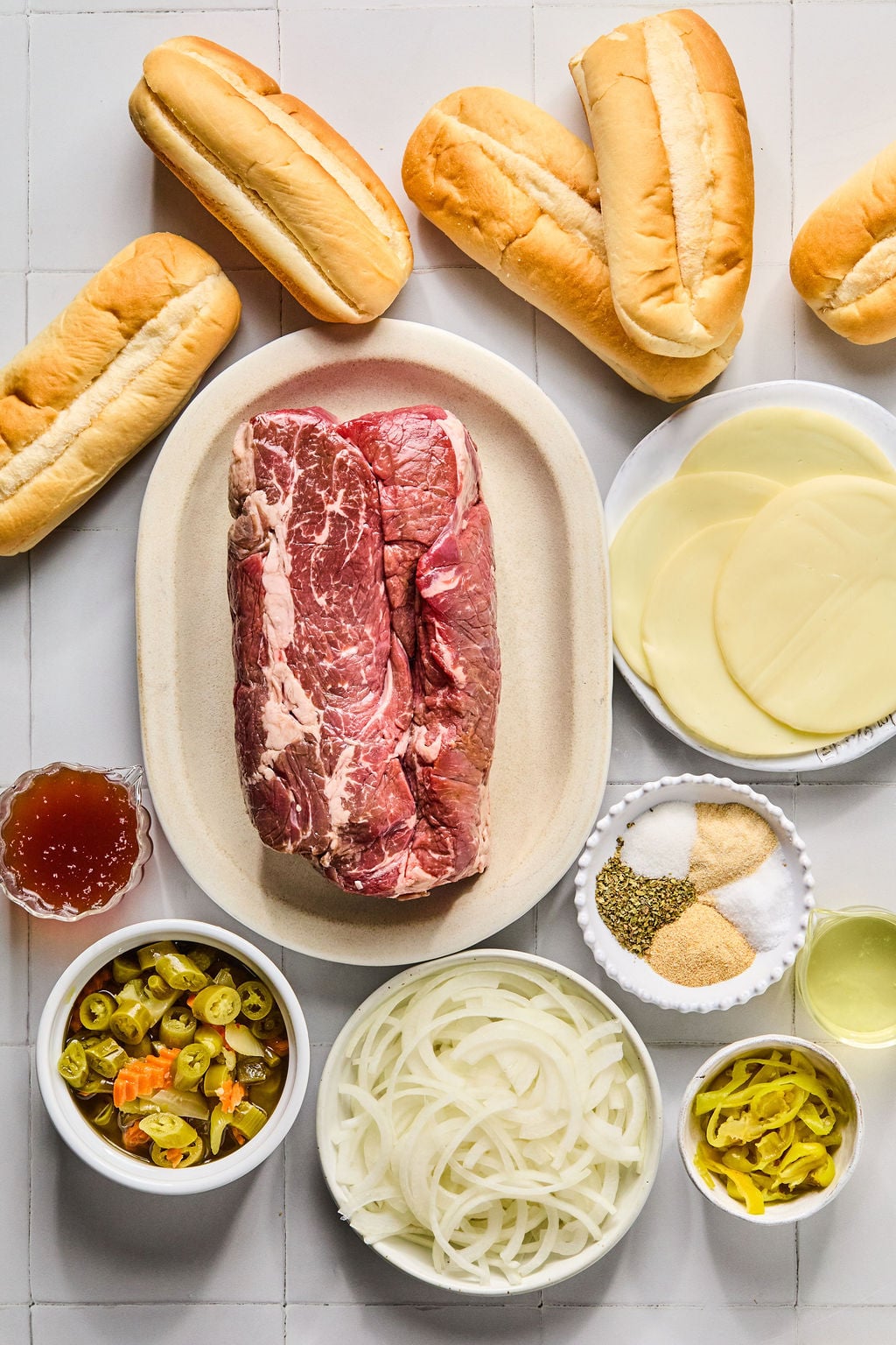 A platter with raw beef, hoagie rolls, provolone cheese slices, sliced onions, pickled peppers, seasoning, and a small bowl of broth arranged on a white surface.