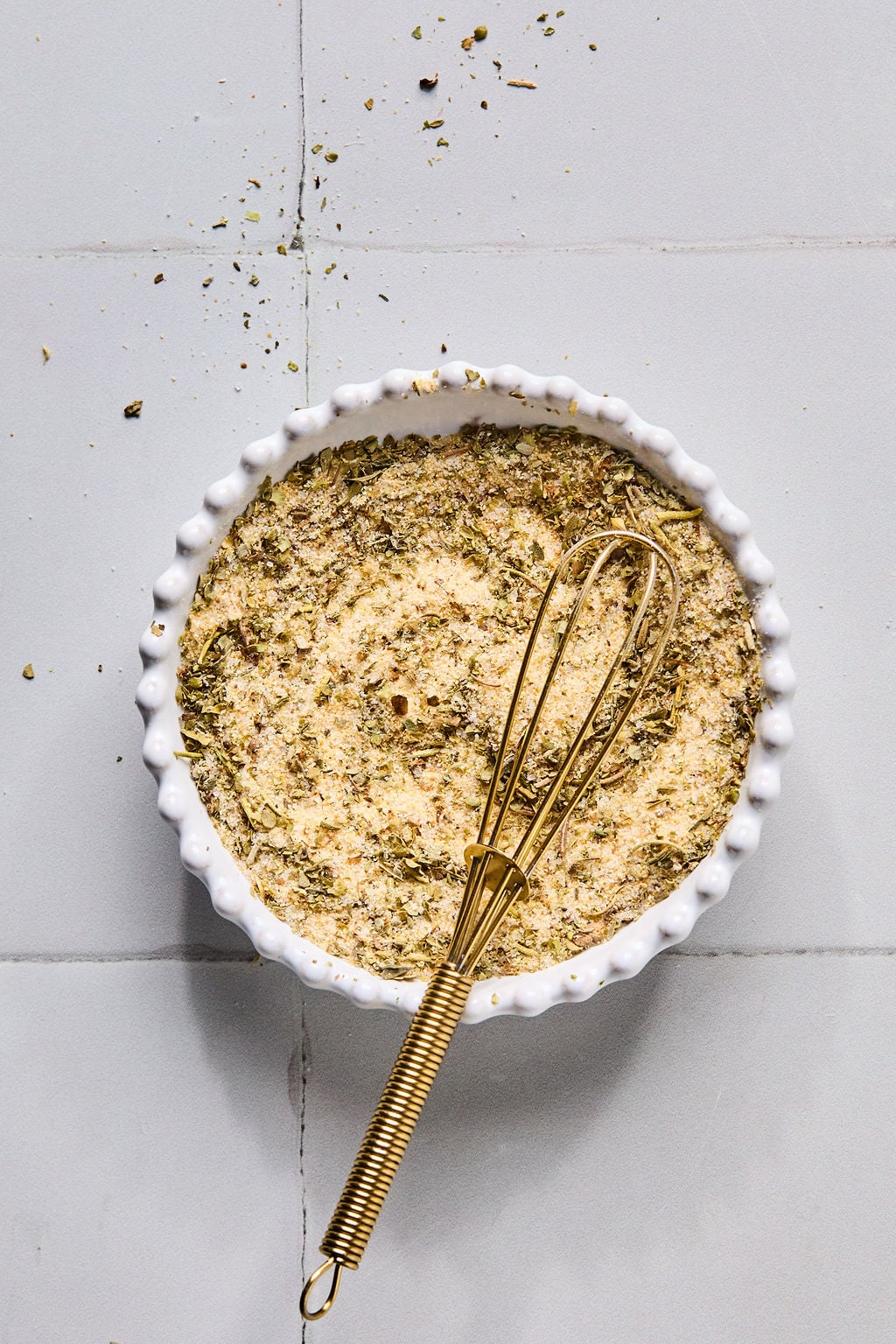 A small white bowl filled with a dry seasoning mix and a gold whisk resting inside, placed on a light gray tiled surface.