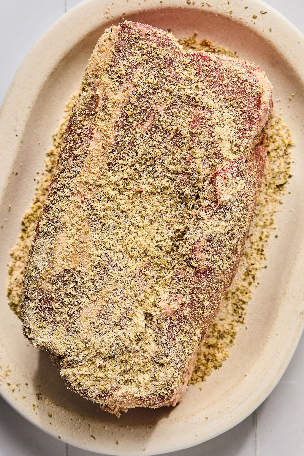 A slab of raw meat covered with a dry seasoning rub sits on an oval beige plate.