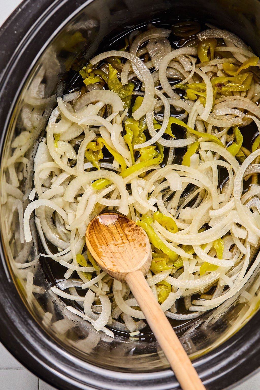 Sliced onions and pepperoncini peppers in a slow cooker with a wooden spoon, prepared for cooking.