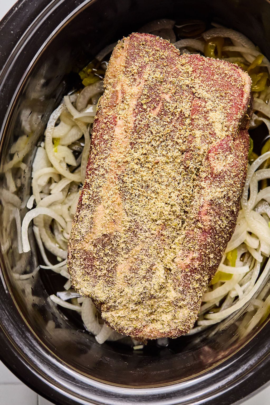 A raw, seasoned beef chuck roast sits on top of sliced onions in a slow cooker, ready to be cooked.