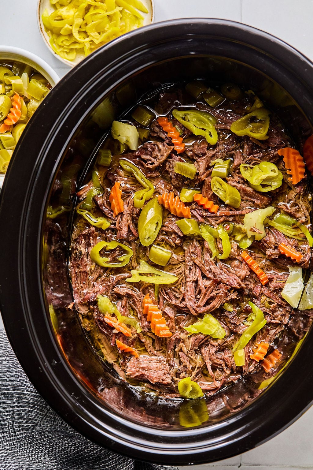 Shredded beef roast with sliced carrots, celery, and pepperoncini peppers in a slow cooker, with small bowls of extra vegetables on the side.