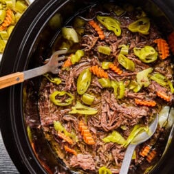 Shredded beef roast with sliced carrots and pepperoncini peppers in a slow cooker, with serving utensils and a bowl of extra peppers nearby.
