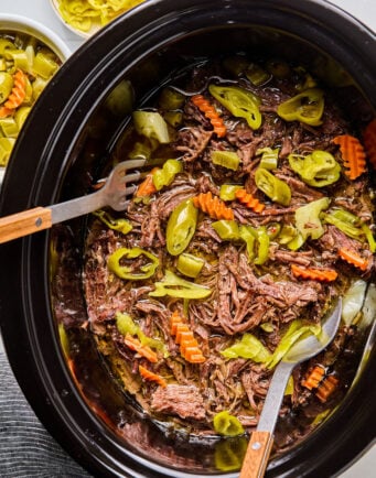Shredded beef roast with sliced carrots and pepperoncini peppers in a slow cooker, with serving utensils and a bowl of extra peppers nearby.