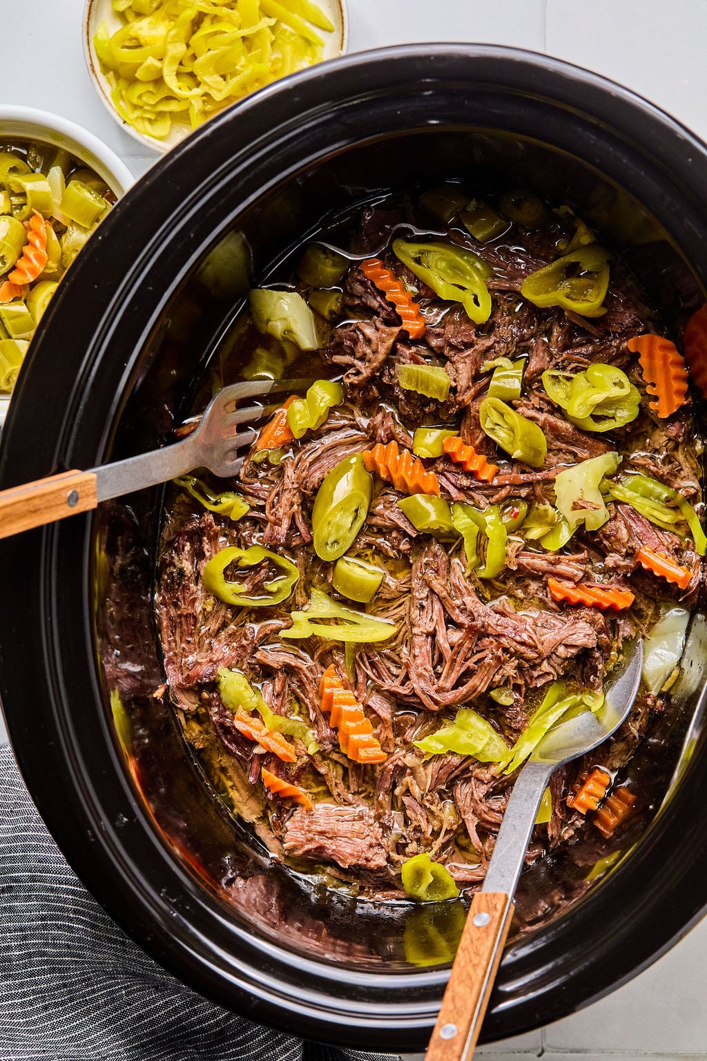 Shredded beef roast with sliced carrots and pepperoncini peppers in a slow cooker, with serving utensils and a bowl of extra peppers nearby.